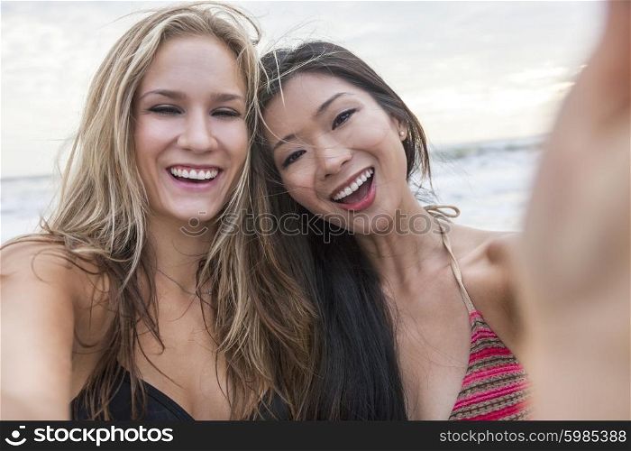 Two young women girls, one Asian Chinese, one blond, wearing bikinis on a beach, laughing taking selfie photograph with digital camera