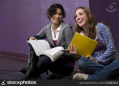 Two young women holding their textbooks and sitting in a corridor