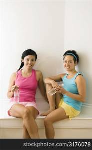 Two young women in fitness clothes holding water bottles sitting smiling.