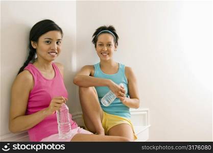 Two young women in fitness clothes holding water bottles sitting smiling.