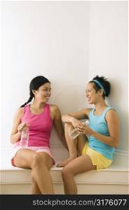 Two young women in fitness clothes holding water bottles sitting smiling and talking.