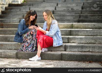 Two young women looking at an smart phone outdoors sitting on urban steps. Friends girls.
