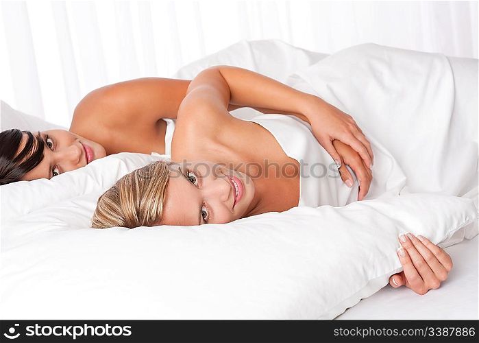 Two young women lying down in white bed looking at camera