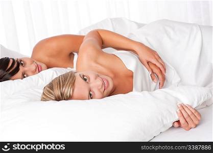 Two young women lying down in white bed looking at camera
