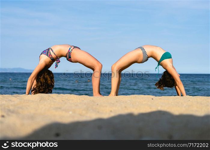 Two young women on the beach practice yoga in bridge position Setu Bandhasana asana by the seaside in sunny day