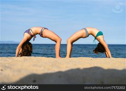 Two young women on the beach practice yoga in bridge position Setu Bandhasana asana by the seaside in sunny day
