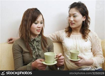 Two young women sitting on a couch holding tea cups