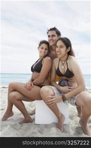 Two young women sitting on an ice box with a young man standing behind them on the beach