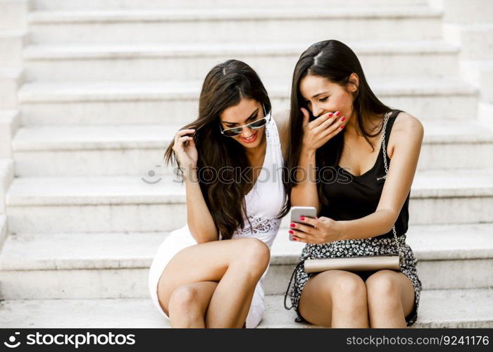 Two young women sitting on the stone steps outside and looking at the phone