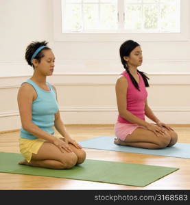 Two young women sitting on yoga mats with eyes closed.