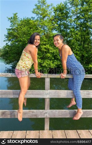 Two young women sitting together on bridge railing at water