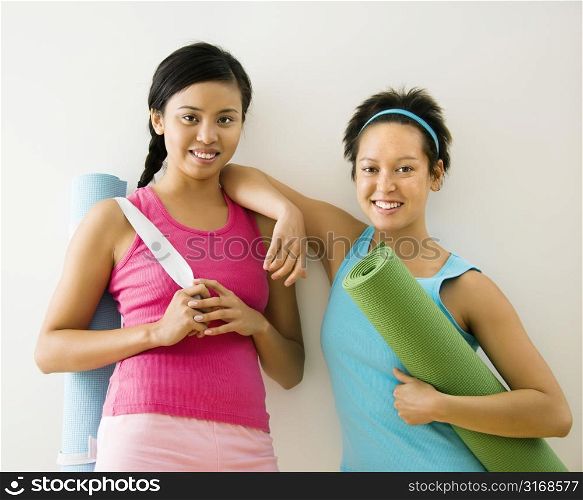 Two young women standing in workout clothes holding yoga mats and smiling.