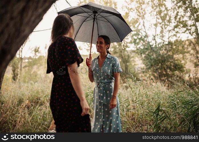 Two young women standing with umbrella on field among trees wearing dresses and sneakers