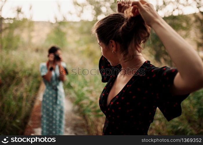 Two young women taking photos each other in the forest among trees wearing dresses
