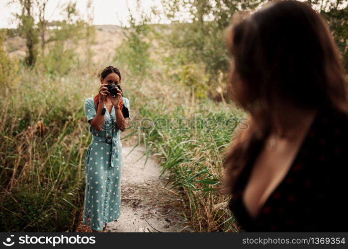 Two young women taking photos each other in the forest among trees wearing dresses