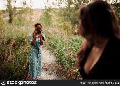 Two young women taking photos each other in the forest among trees wearing dresses