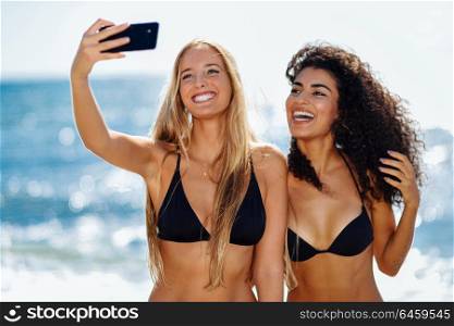 Two young women taking selfie photograph with smart phone in swimwear on a tropical beach. Funny caucasian and arabic females wearing black bikini.