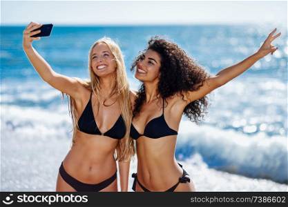 Two young women taking selfie photograph with smart phone in swimwear on a tropical beach. Funny caucasian and arabic females wearing black bikini.. Two women taking selfie photograph with smartphone in the beach