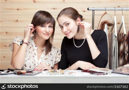 Two young women trying on earrings on wooden background. Clothes, cosmetic and accessories around them