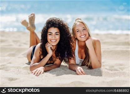 Two young women with beautiful bodies in swimsuit on a tropical . Two young women with beautiful bodies in swimwear on a tropical beach. Funny caucasian and arabic females wearing black and white swimsuits lying on the sand on the beach
