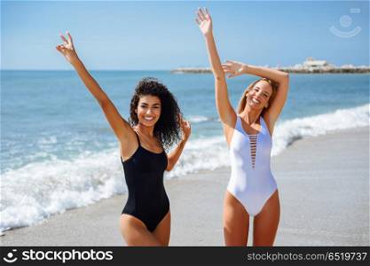 Two young women with beautiful bodies in swimsuit on a tropical . Two young women with beautiful bodies in swimwear on a tropical beach. Funny caucasian and arabic females wearing black and white swimsuits waving with their hands
