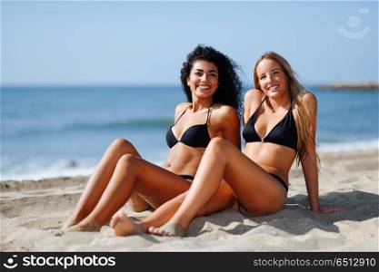 Two young women with beautiful bodies in swimwear on a tropical . Two young women with beautiful bodies in swimwear on a tropical beach. Funny caucasian and arabic females wearing black bikini sitting on sand.
