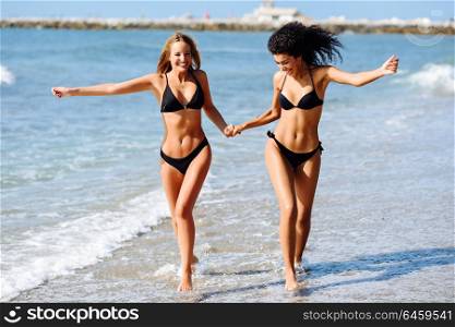 Two young women with beautiful bodies in swimwear on a tropical beach. Funny caucasian and arabic females wearing black bikini walking along the shore holding hands.