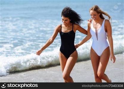 Two young women with beautiful bodies in swimwear on a tropical beach. Funny caucasian and arabic females wearing black and white swimsuits walking along the shore.. Two young women with beautiful bodies in swimsuit on a tropical beach