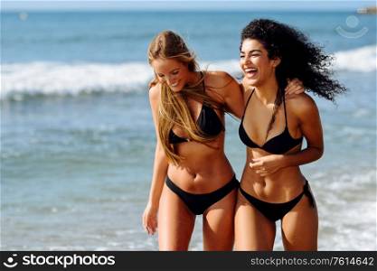 Two young women with beautiful bodies in swimwear on a tropical beach. Funny caucasian and arabic females wearing black bikini walking along the shore.. Two young women with beautiful bodies in swimwear on a tropical beach
