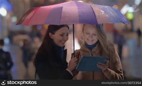 Two young women with tablet computer in rainy evening in the city. They standing under colorful umbrella and having exctied talk while looking through the photos or browsing the internet