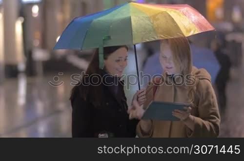 Two young women with umbrella stand talking and gesticulating cheerfully on the street on rainy day, they watch something on electronic tablet