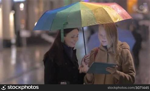 Two young women with umbrella stand talking and gesticulating cheerfully on the street on rainy day, they watch something on electronic tablet