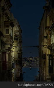 Typical narrow street leading to the sea on the island of Malta at night. Buildings with traditional colorful maltese balconies in historical part of Valletta.