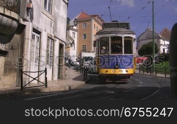 Um die Kurve bergabfahrende blau-gelbe Tram in Lissabon;dahinter fahren Autos unnd LieferwSgen.