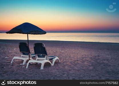 Umbrella and two sunbeds on beach at sunset. Sandy beach with sunbeds against the backdrop of the sea and sunset sky. The concept of relaxation and travel. Umbrella and two sunbeds on beach at sunset
