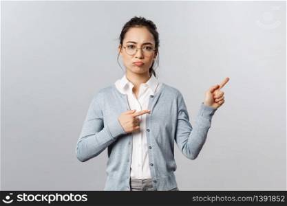 Unamused, gloomy and complaining young asian girl in glasses, sulking upset, grimacing unsatisfied and pointing fingers right with regret and disappointment, standing grey background.. Unamused, gloomy and complaining young asian girl in glasses, sulking upset, grimacing unsatisfied and pointing fingers right with regret and disappointment, standing grey background