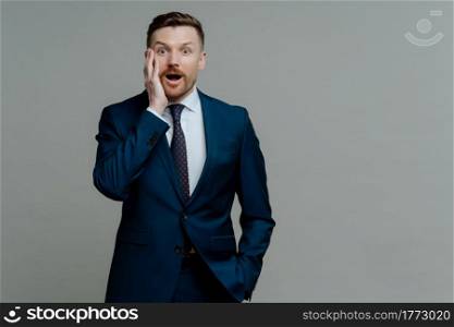 Unbelievable news. Young shocked businessman in suit looking at camera with surprised face expression and opened mouth while standing against grey background. Business people and emotions concept. Surprised businessman in suit looking at camera with shocked face expression