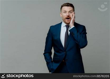 Unbelievable news. Young shocked businessman in suit looking at camera with surprised face expression and opened mouth while standing against grey background. Business people and emotions concept. Surprised businessman in suit looking at camera with shocked face expression