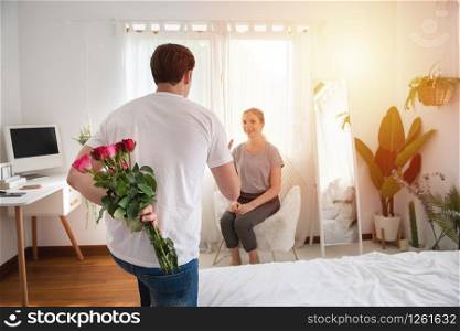 Unexpected moment in routine everyday life! photo of man?s hands hiding holding chic bouquet of red roses with behind back, happy woman is on blurred background with copy space