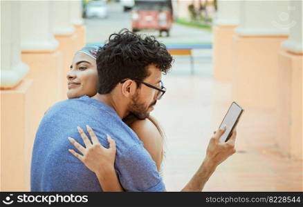 Unfaithful man looking at the cell phone while hugging his girlfriend outdoors. Unfaithful boyfriend hugging his girlfriend and looking at the cell phone. Concept of unfaithful man using cell phone