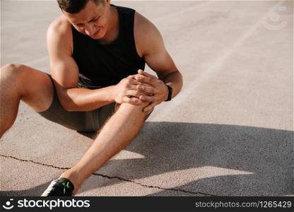 Unfortunate side of sports. Young handsome athlete sitting on the racetrack holding on to his injured knee. close up of runner touching painful twisted or broken ankle shin or calf athlete runner training accident sport running ankle sprain concept