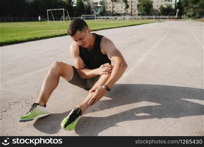 Unfortunate side of sports. Young handsome athlete sitting on the racetrack holding on to his injured knee. close up of runner touching painful twisted or broken ankle shin or calf athlete runner training accident sport running ankle sprain concept