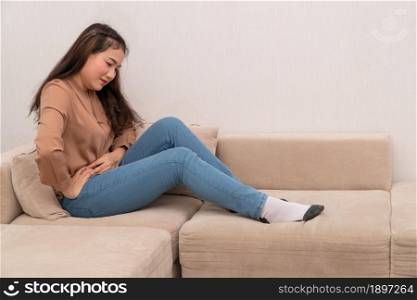 Unhappy Asian woman sitting on sofa and holding on stomach suffering from menstruation pain