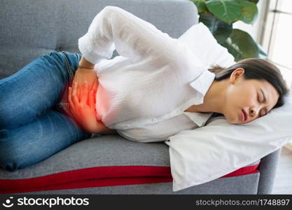 Unhappy Asian woman sitting on the sofa and holding on stomach suffering. Abdominal pain that comes from menstruation, diarrhea, or indigestion. Sickness and healthcare concept