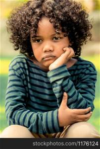 Unhappy bored little african american kid sitting in the park. The boy showing negative emotion. Child trouble concept.