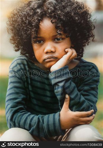 Unhappy bored little african american kid sitting in the park. The boy showing negative emotion. Child trouble concept.. Unhappy little african american kid in the park.