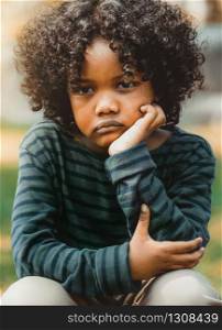 Unhappy bored little african american kid sitting in the park. The boy showing negative emotion. Child trouble concept.. Unhappy little african american kid in the park.