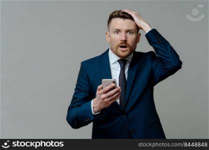 Unhappy businessman in suit holding cellphone and getting bad unpleasant news, looking at camera with open mouth and shocked face expression while standing against grey background. Failure concept. Shocked businessman in suit feeling upset while receiving unexpected news