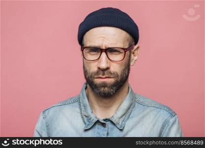 Unhappy discontent unshaven male looks with grumpy expression into camera, wears glasses black hat and stylish denim shirt, isolated over pink background. Displeased middle aged man poses indoor