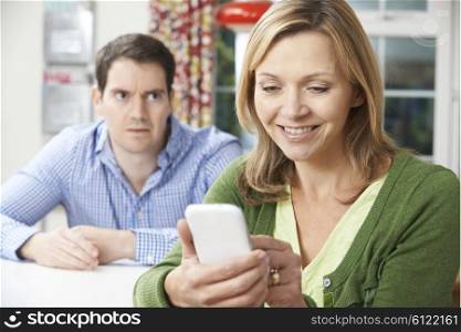 Unhappy Man Sitting At Table As Partner Texts On Mobile Phone